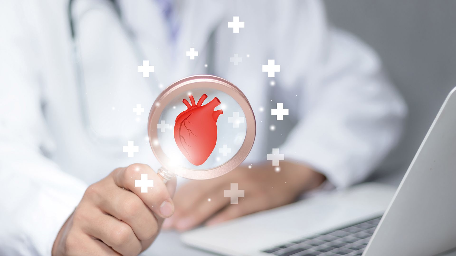 Arzt am Laptop, mit einer Lupe in der Hand, in der ein anatomisches Herz abgebildet ist