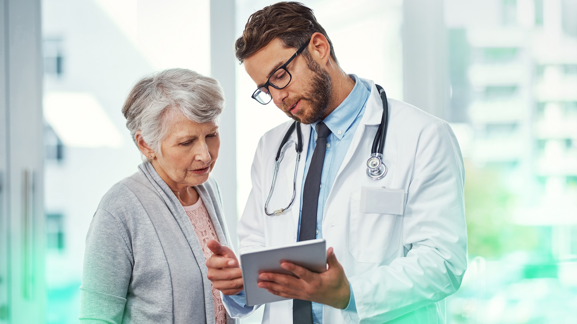 Bild, Arzt mittleres Alter hält Tablet in der Hand, erklärt älterer Patientin links neben ihm etwas