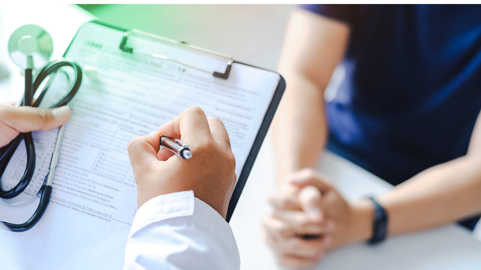Es ist eine Hand einer Person zu sehen, die einen Stift hält und ein Formular auf einem Klemmbrett ausfüllt. In der anderen Hand hält die Person ein Stethoskop.