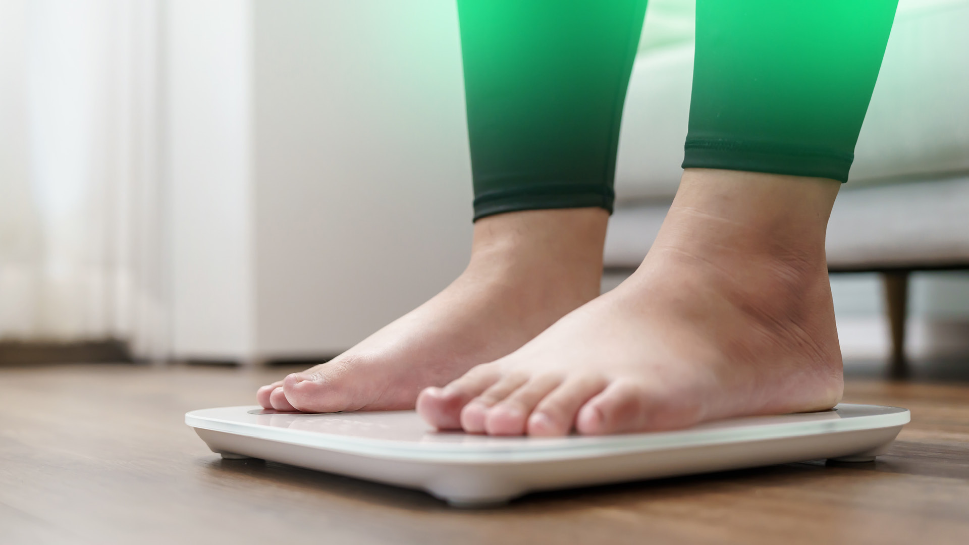 Nackte Füße stehen auf einer weißen Waage, vermutlich weiblich, trägt Leggings, im Hintergrund Bett
