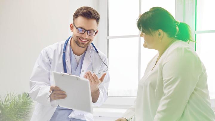 Freundlicher Arzt mit Stethoskop erklärt einer Patientin in heller Praxis die Ergebnisse auf einem Klemmbrett – medizinische Beratung, Gesundheitscheck und Arzt-Patienten-Gespräch in moderner Arztpraxis.