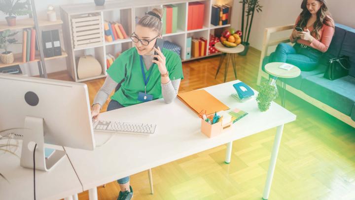 Medizinische Fachangestellte als Coach für chronische Patienten arbeitet am Computer in einer hellen Praxis und berät eine Patientin im Wartebereich.